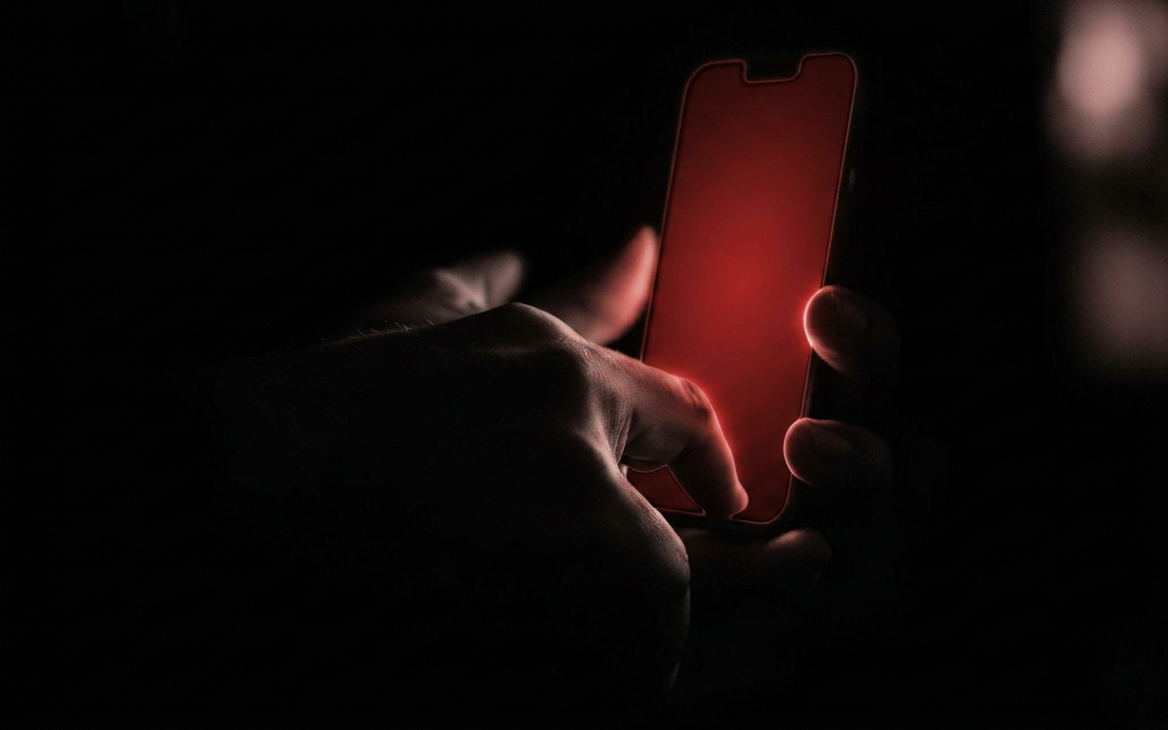 Editorial-Foto im Dunkeln: Eine Hand hält ein Smartphone mit leuchtend rotem Display, nur das rote Licht und die Finger sind sichtbar