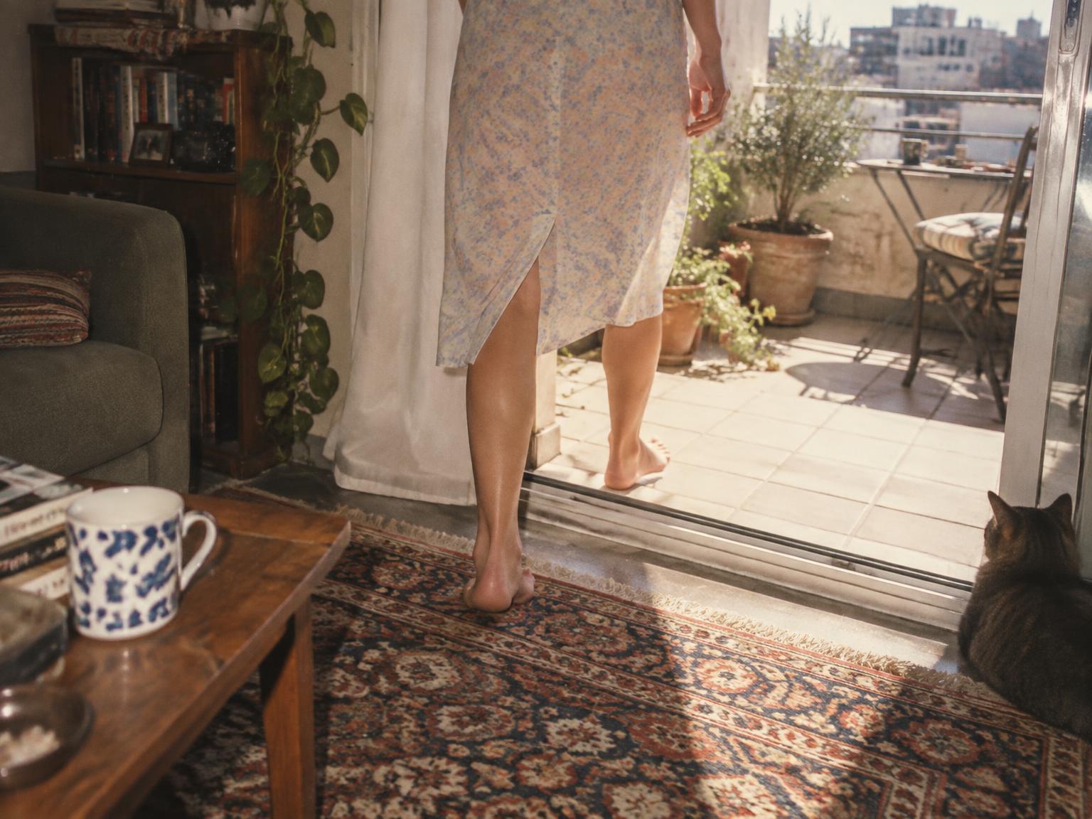 Analoger Schnappschuss einer Frau, die barfuß vom Wohnzimmerteppich auf einen sonnigen Balkon tritt.