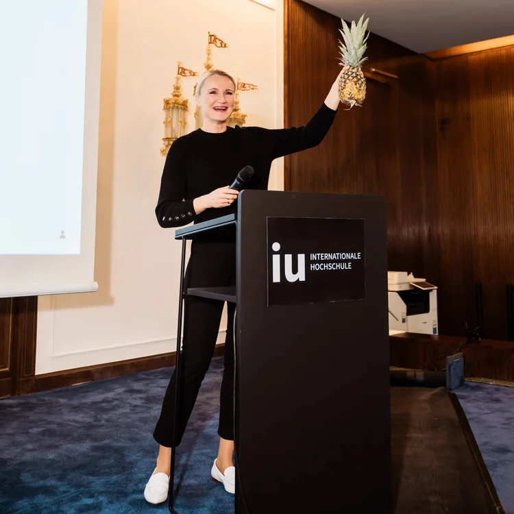 Henriette Hochstein-Frädrich lachend am IU-Rednerpult mit Ananas in der Hand · Keynote „Human & AI“ beim AI BarCamp der IU Internationalen Hochschule 2025 in Köln