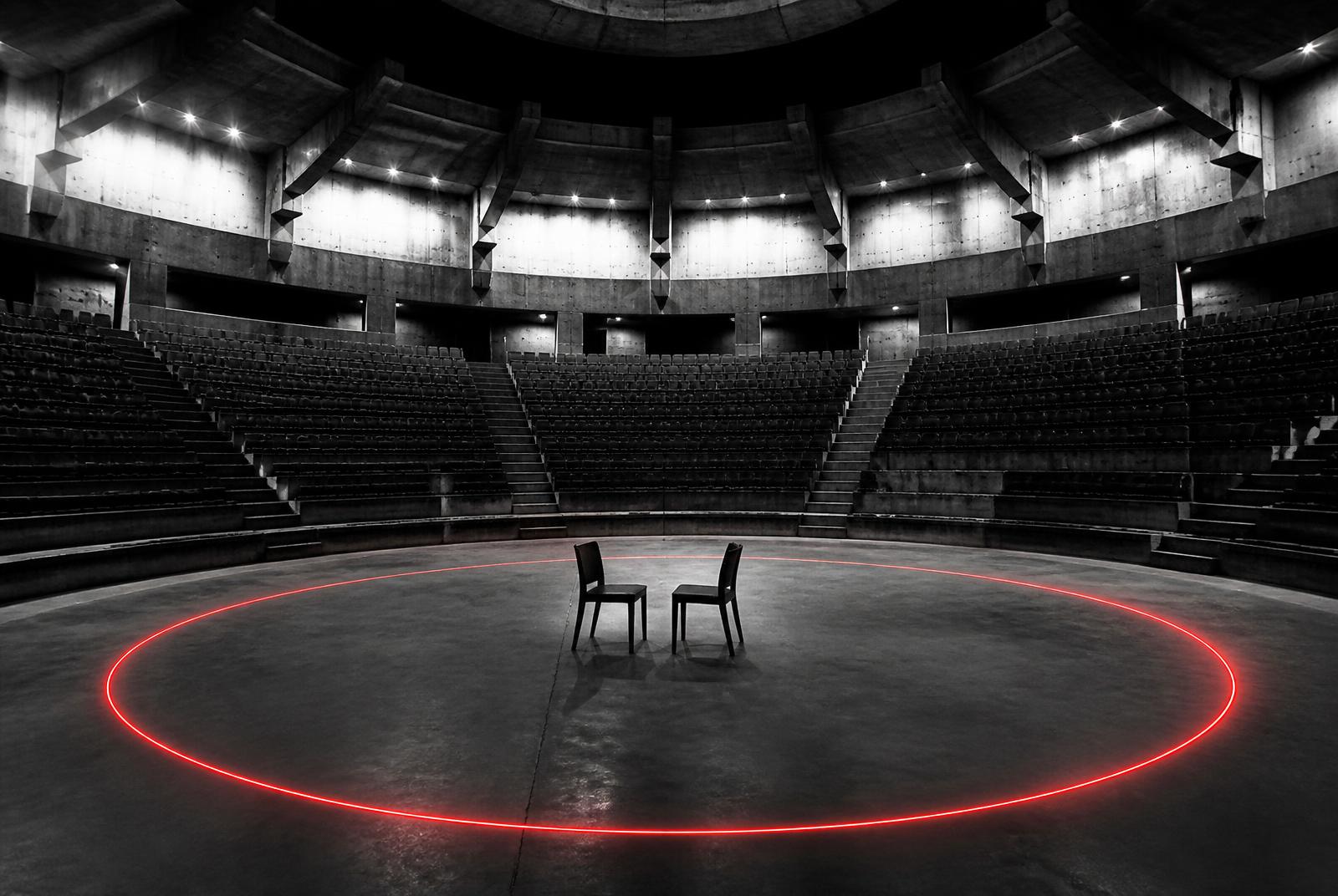 Editorial-Schwarz-Weiß-Foto mit einem einzigen roten Akzent: eine riesige leere Beton-Arena mit hunderten leeren Sitzreihen, in deren Mitte nur zwei einfache Stühle einander zugewandt stehen; ein dünner roter Neonkreis auf dem Boden ist die einzige Farbe im Bild.