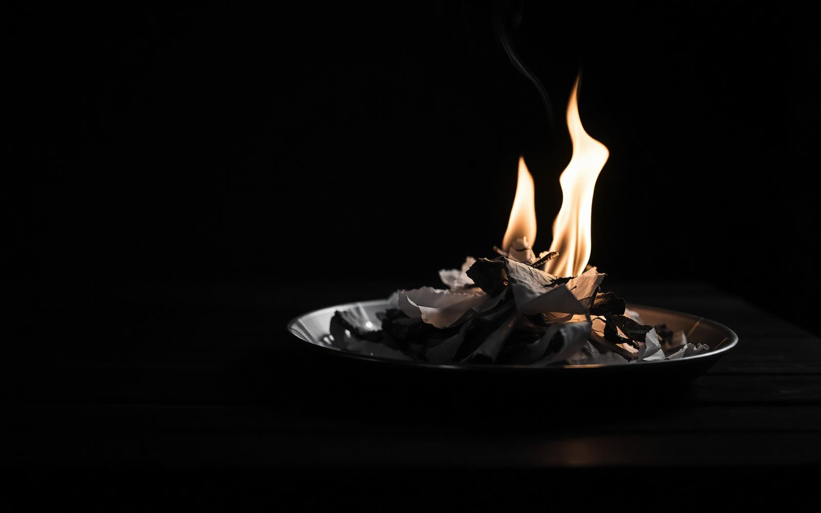 Cinematisches Schwarz-Weiß-Editorial-Stillleben: ein kleiner Haufen zerrissener Papierschnipsel brennt auf einem Backblech auf einem dunklen Terrassentisch bei Nacht — warm-goldene Flammen als einzige Farbe im monochromen Bild, Metapher für Loslassen, Alchemie und ein Feuerritual zum Jahreswechsel.
