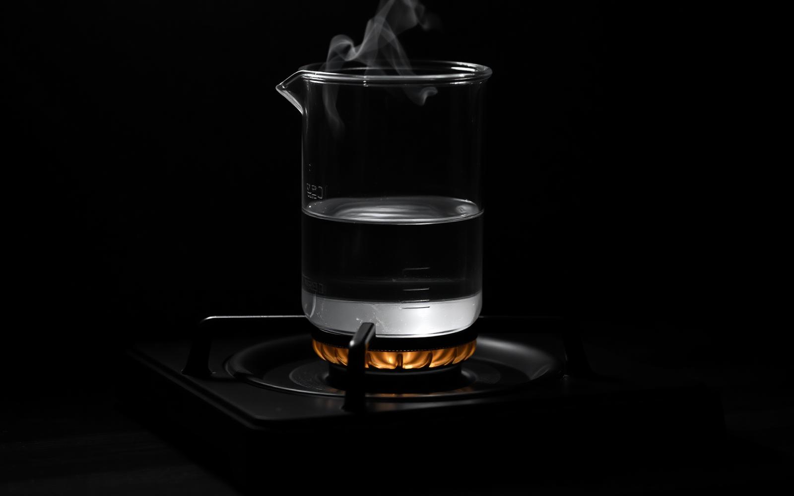 Cinematisches Schwarz-Weiß-Editorial-Stillleben auf dunklem Holz: ein Glasbecher voller Wasser auf einer Herdplatte, feiner Dampf steigt auf, eine warm-goldene Flamme darunter als einzige Farbe im monochromen Bild — Metapher für den Frosch im heißen Wasser und Shifting Baselines.