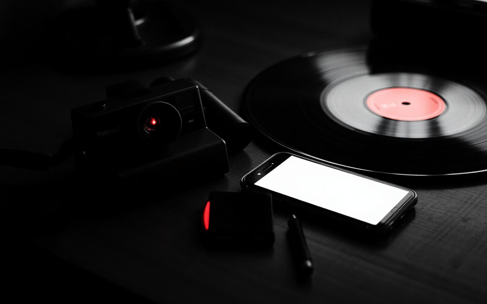 Editorial-Schwarz-Weiß-Foto mit rotem Akzent: Polaroid-Kamera und Vinylplatte auf einem Holztisch neben einem leuchtenden Smartphone, das rote Label der Platte ist die einzige Farbe im Bild
