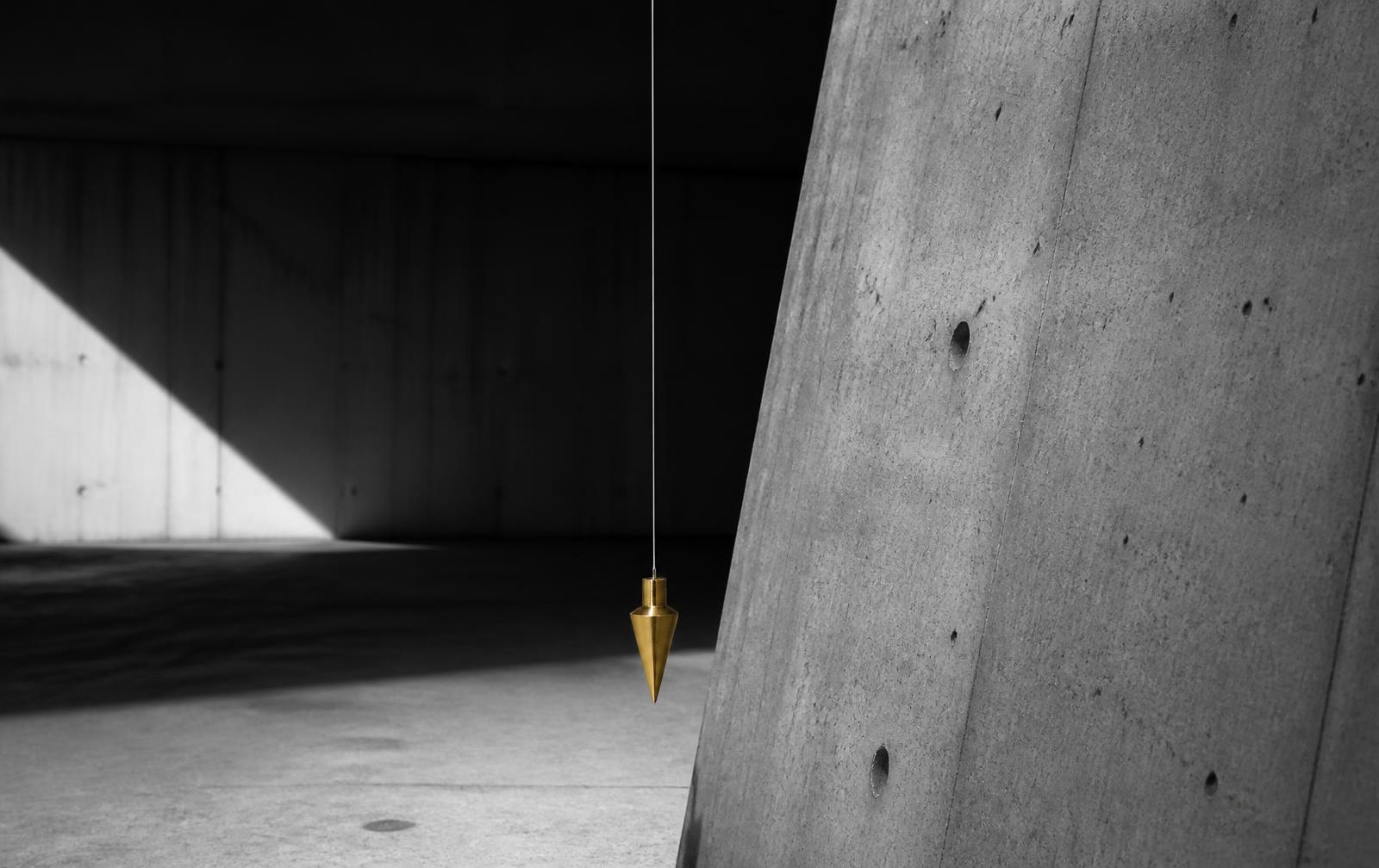 Modernes Schwarz-Weiß-Editorial-Foto in einer rohen Betonhalle mit hartem Licht: an einem dünnen Stahldraht hängt ein präzise gedrehtes, warm-goldenes Senklot vollkommen ruhig vor einer leicht geneigten Betonwand. Das Gold ist die einzige Farbe im monochromen Bild — Metapher für das innere Lot, Balance und rechtzeitiges Gegensteuern.
